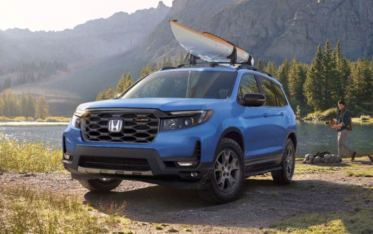 Vista delantera de 3/4 desde el lado del conductor de la Honda Passport TrailSport 2025 en Diffused Sky Blue Pearl estacionado en un campamento junto a un lago