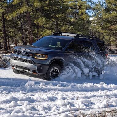 2027 Honda Passport TrailSport Elite in Smoke Blue Pearl front driving through deep snow in a pine forest (Mid-Size SUV).
