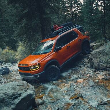 2027 Honda Passport TrailSport Elite in Sunset Orange profile driving through a rocky stream in the forest (Mid-Size SUV).